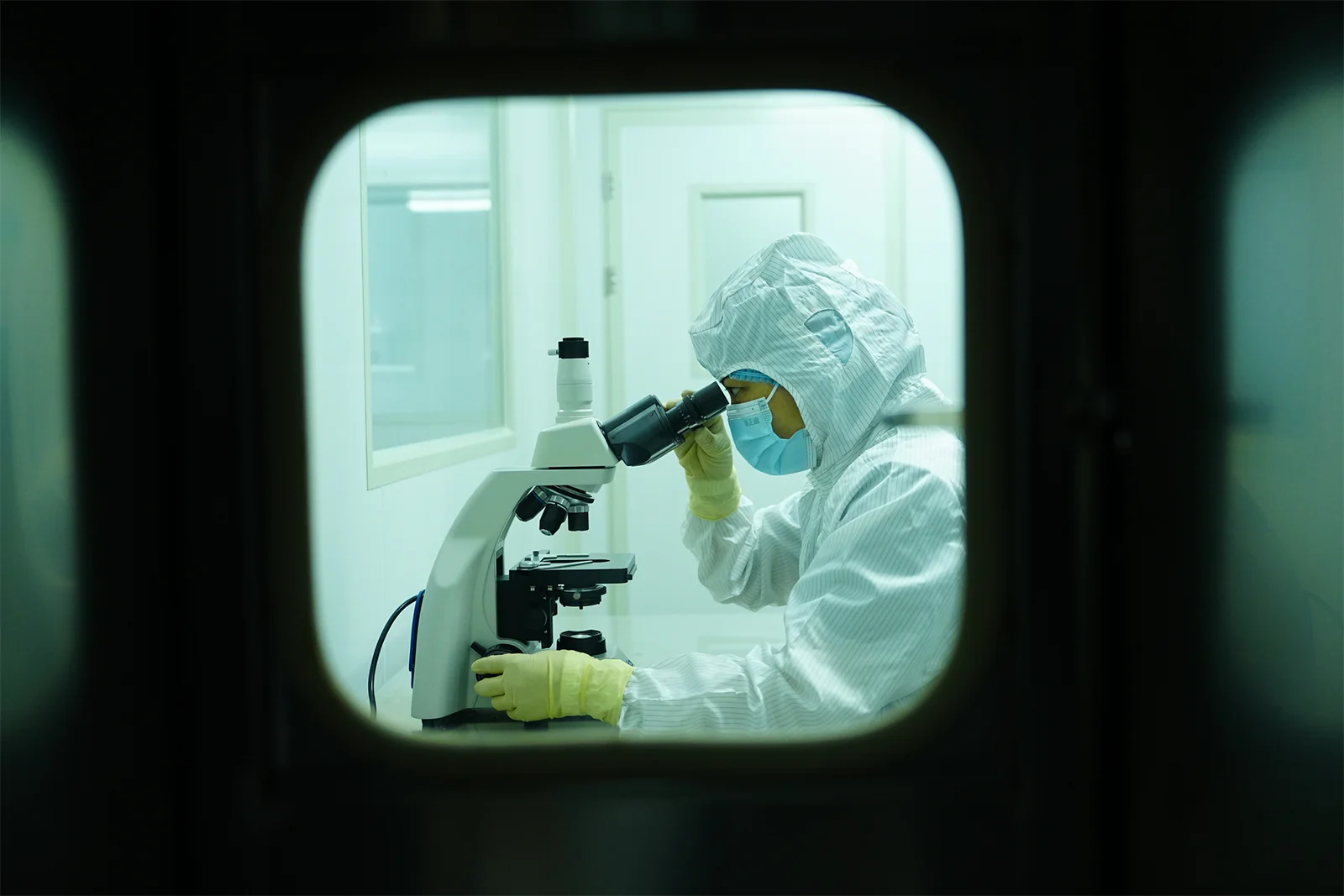 View through a cleanroom observation window of a Baird Medical specialist examining a sample under a microscope