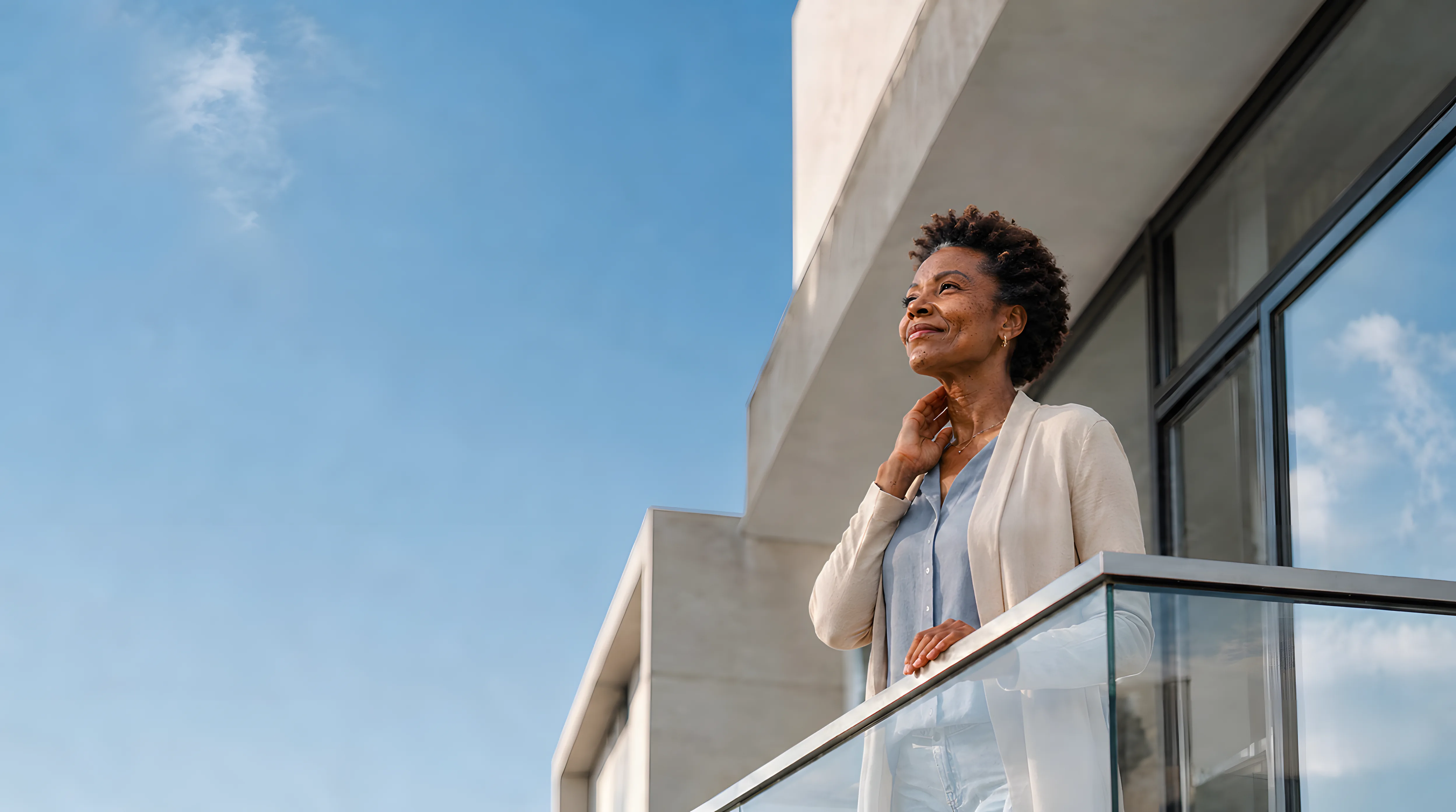 A woman stands on a sunlit balcony looking toward an open sky, one hand resting near her neck